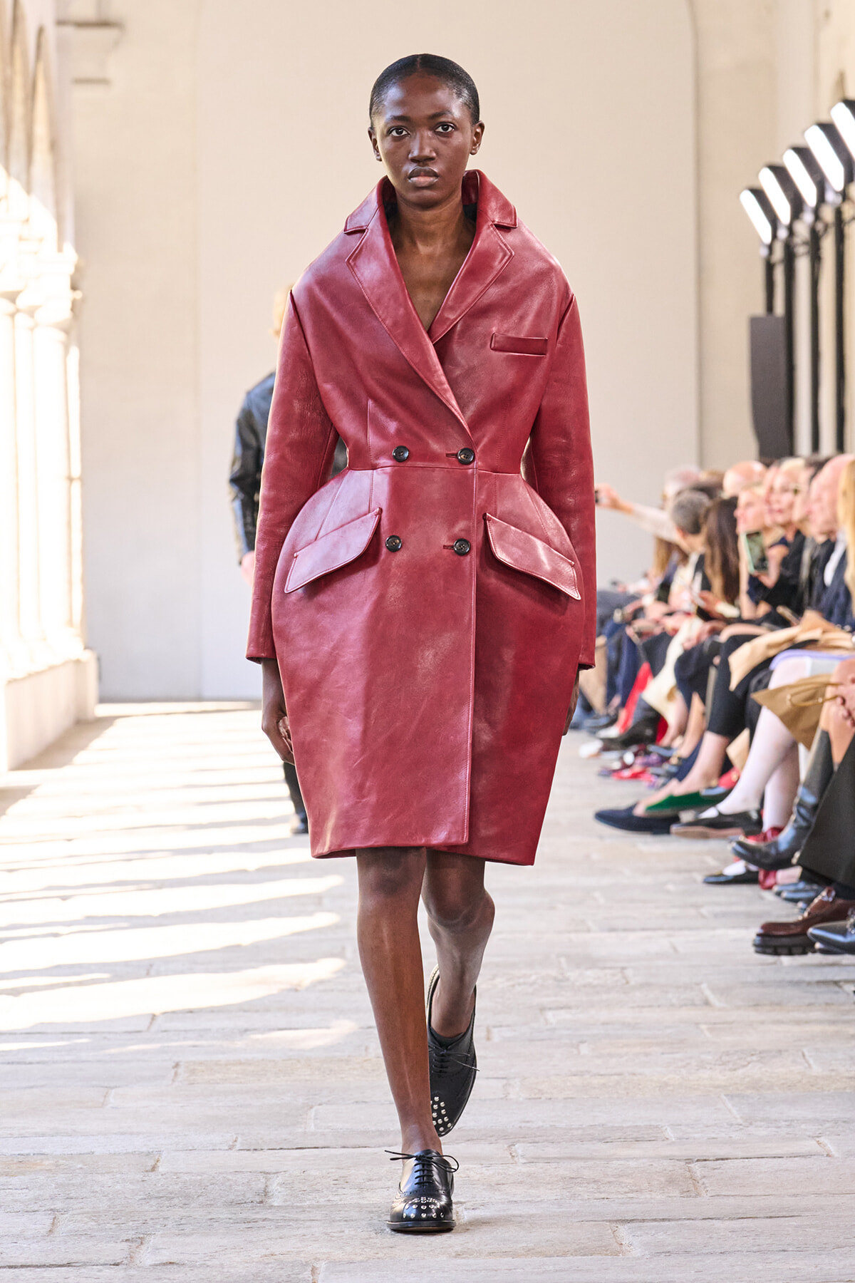 Model walks the runway wearing a burgundy leather double-breasted trench with oversized pockets and a belted waist in an outdoor-style show.