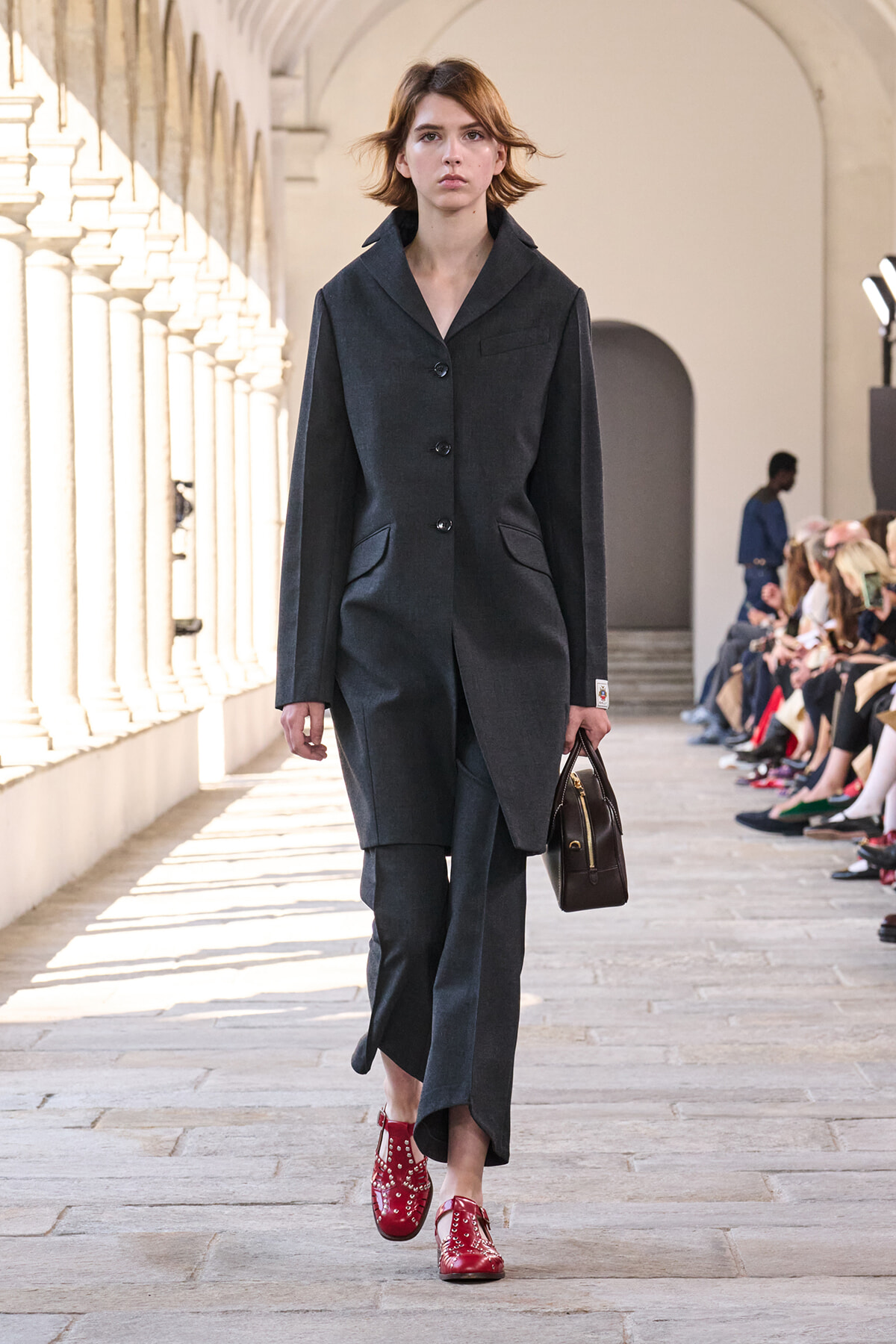 Model walking a fashion runway in a charcoal gray suit, red patent leather shoes, and a small brown handbag.