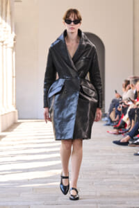 Model walking the runway in a black leather belted trench coat and sunglasses, neutral stance, arched backdrop.