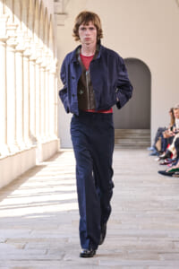 Male model walks a runway in a navy outfit: blue jacket, matching trousers, red mesh shirt, brown vest, with audience on the side.
