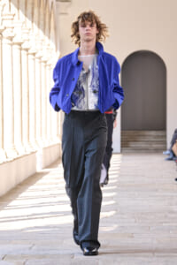Model walking runway in a bright cobalt blue jacket over a metallic silver top and loose charcoal trousers.