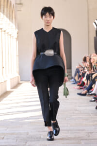 Model walking a runway in a black sleeveless top and pants with a large belt and a green handbag on the catwalk.