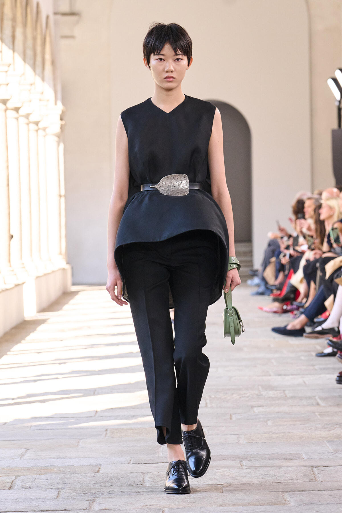 Model walking a runway in a black sleeveless top and pants with a large belt and a green handbag on the catwalk.