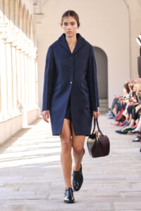 Model walks a runway in a navy coat with a two-tone beltless silhouette, carrying a small dark brown handbag on a stone walkway.