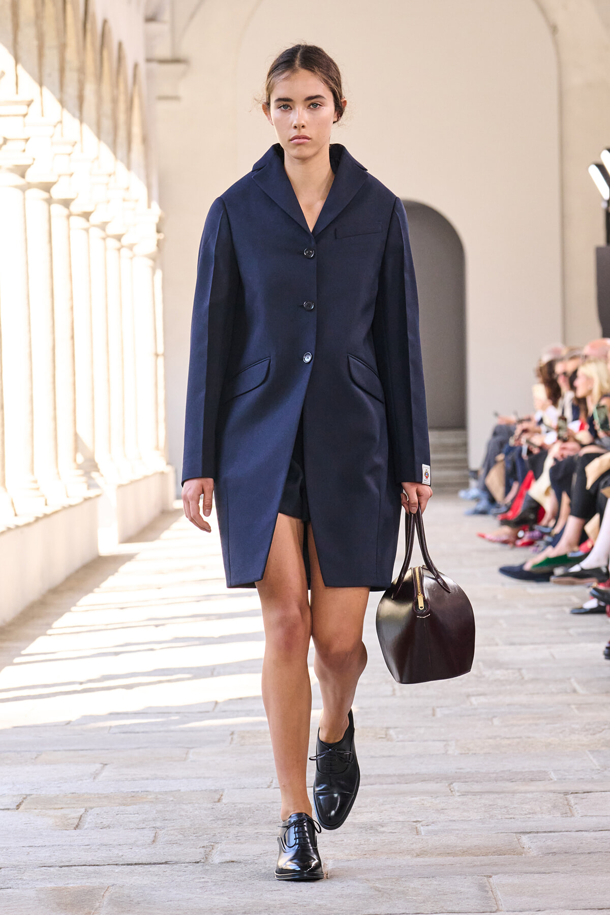 Model walks a runway in a navy coat with a two-tone beltless silhouette, carrying a small dark brown handbag on a stone walkway.