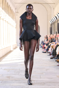 Black female model walks a runway in a structured black peplum dress with studded shoes, audience along the sides in a bright hall.