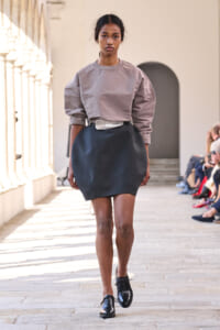 Model walking a fashion runway in a taupe oversized sweatshirt tucked into a dark mini skirt with a metallic belt.