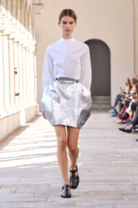Fashion model in a white high-neck blouse and metallic silver bubble skirt walking the runway, with seated audience along the catwalk.
