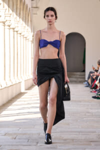 Model walks a runway in a blue twisted bandeau top and black wrap skirt, carrying a small black handbag amid colonnade architecture.