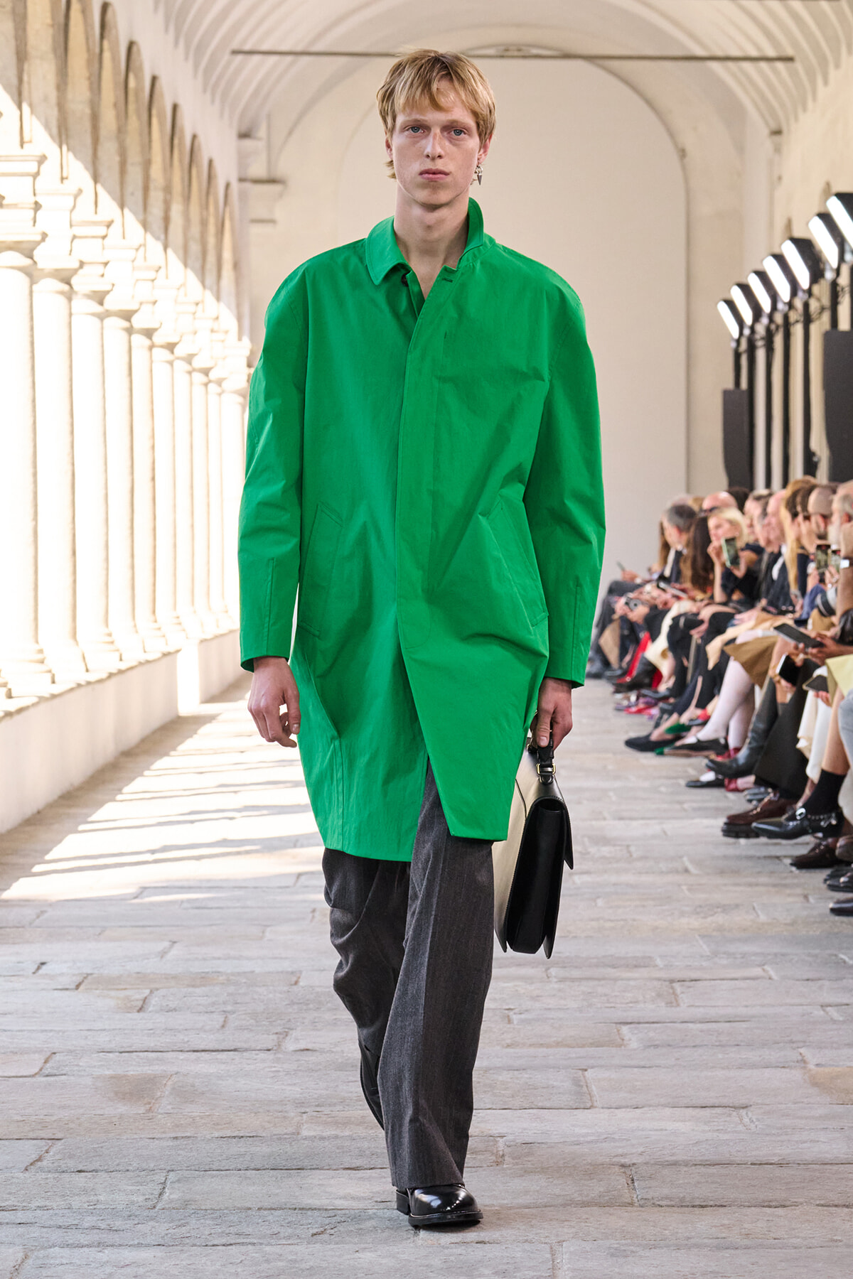 Model walking a fashion runway in a bright green long coat, gray trousers, black shoes, and holding a black handbag.