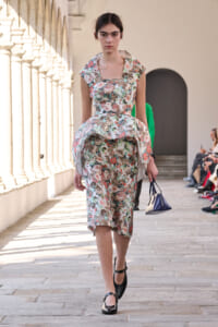 Model walking a floral print dress down a sunlit architectural runway, carrying a small blue handbag and wearing black Mary Jane shoes.