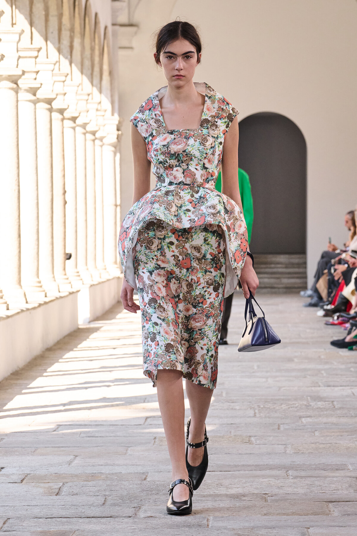 Model walking a floral print dress down a sunlit architectural runway, carrying a small blue handbag and wearing black Mary Jane shoes.