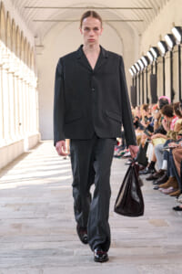 Model walking a runway in a dark grey suit, carrying a large dark burgundy leather bag, with spectators seated along the side.