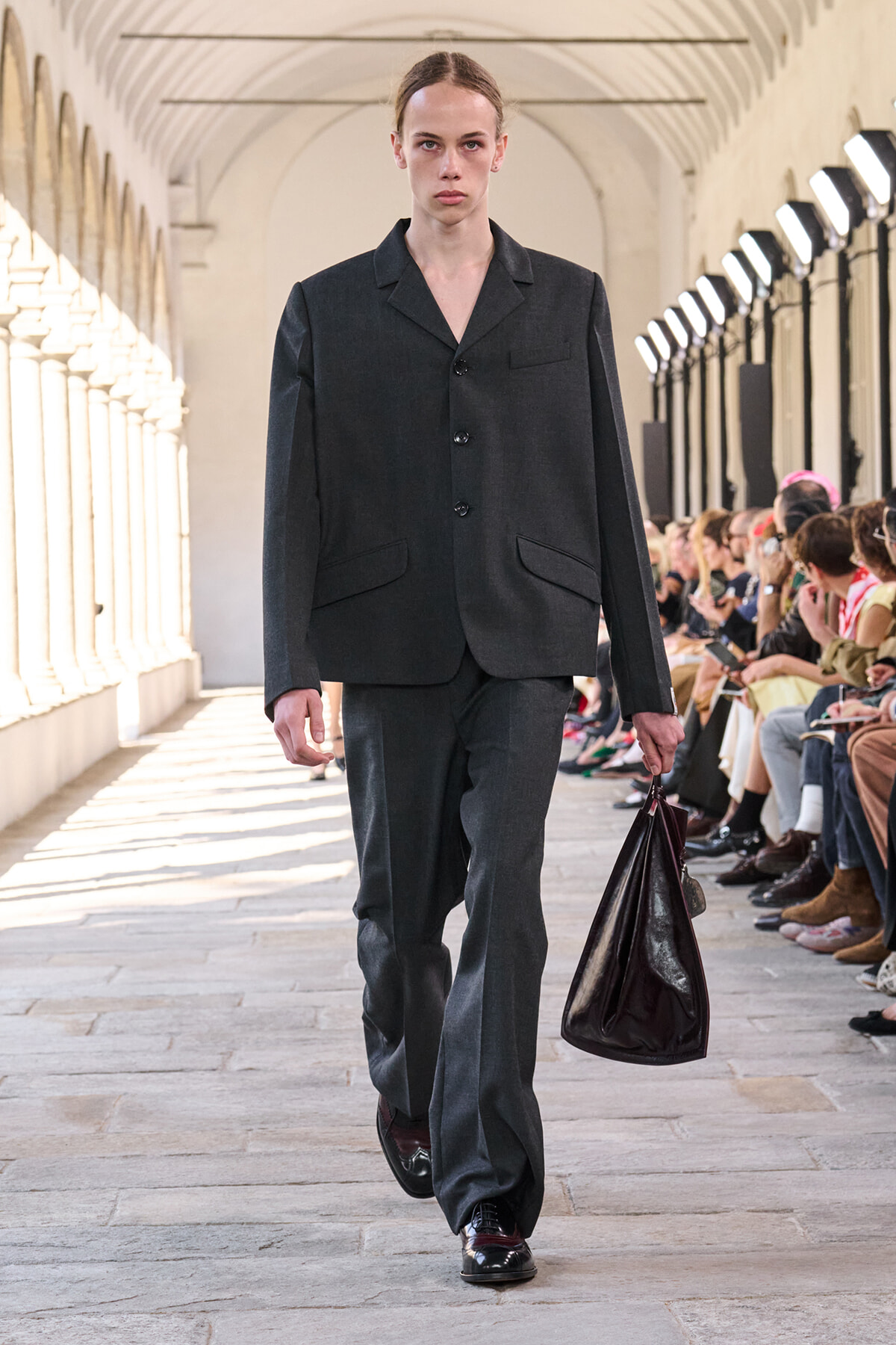 Model walking a runway in a dark grey suit, carrying a large dark burgundy leather bag, with spectators seated along the side.