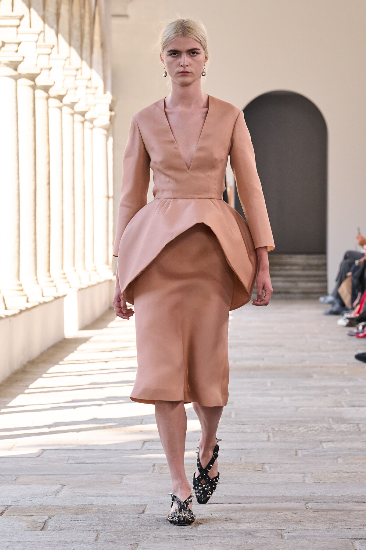 Model walks a peach-pink satin dress with a structured bodice and sculpted peplum on a sunlit runway.