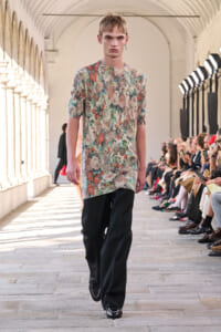 Male model in a colorful floral tunic and black pants walks a fashion runway while seated audience looks on.