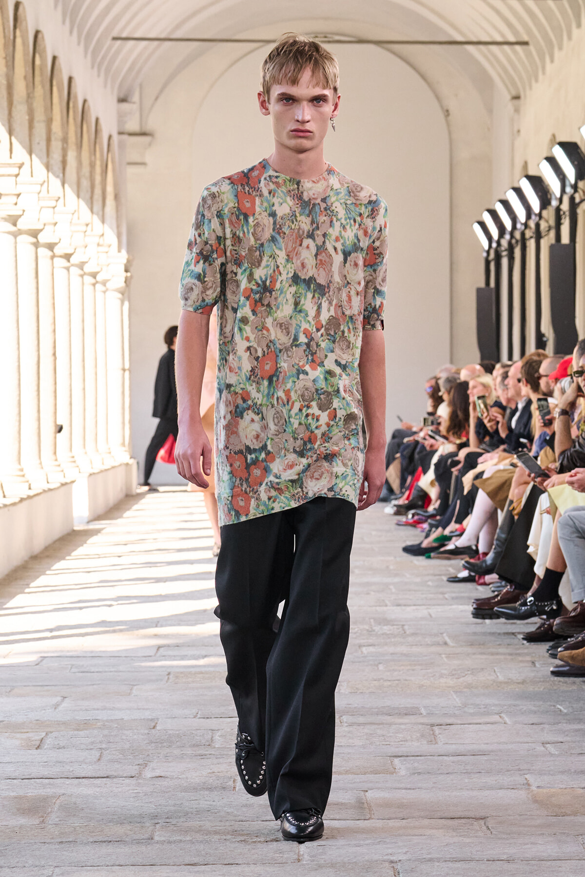 Male model in a colorful floral tunic and black pants walks a fashion runway while seated audience looks on.