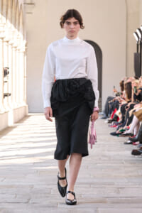 Model walking a fashion show in a white high-neck blouse and black knee-length skirt with a large black fabric rosette at the waist, carrying a pink handbag; audience on either side.
