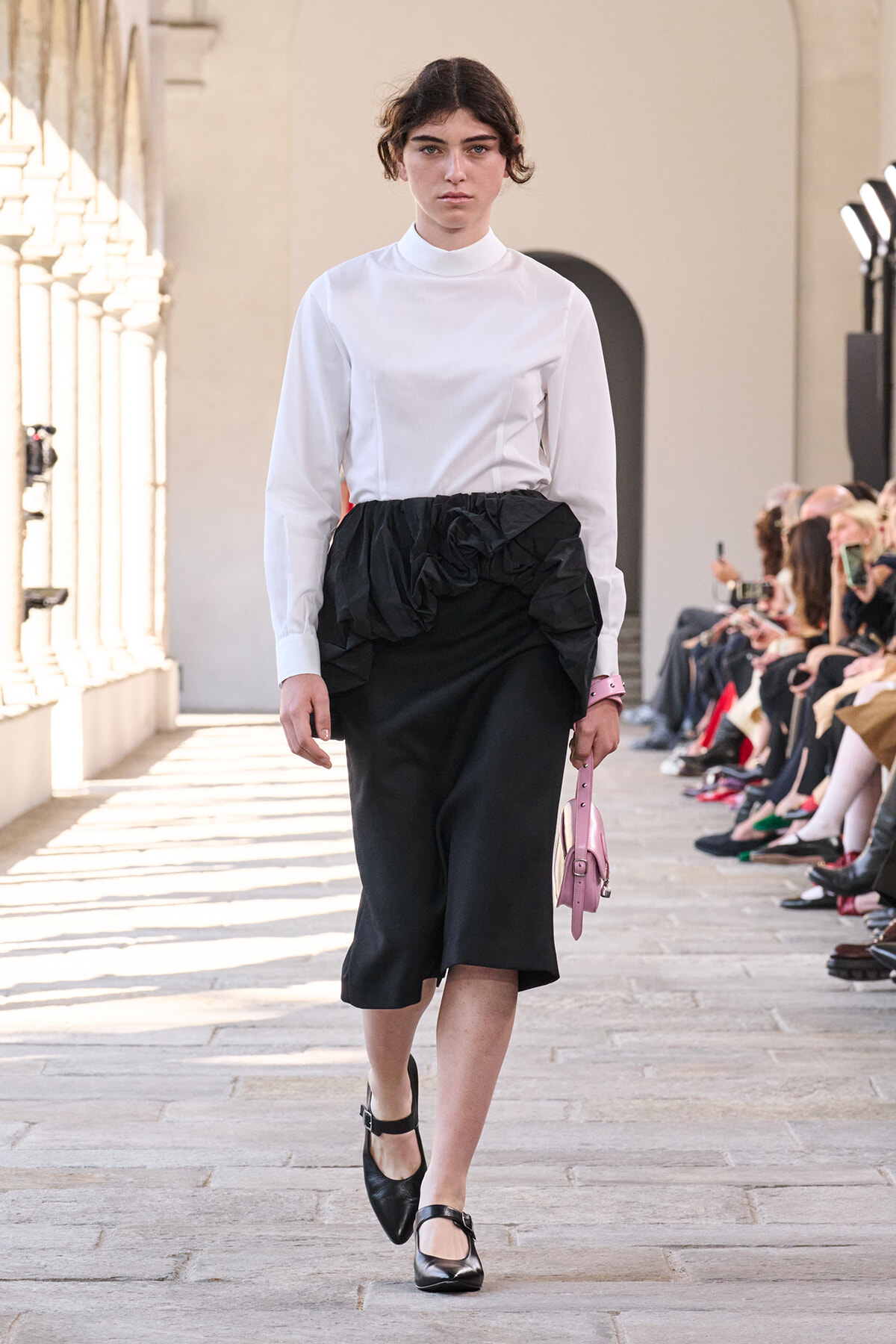 Model walking a fashion show in a white high-neck blouse and black knee-length skirt with a large black fabric rosette at the waist, carrying a pink handbag; audience on either side.