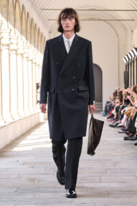 Model walking a runway in a long dark double-breasted coat, black trousers, and a dark leather portfolio bag.
