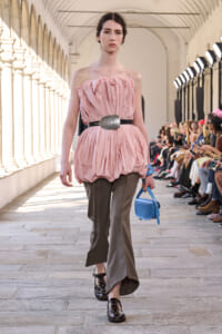 Model walks the runway in a pink, ruched strapless top with a wide metallic belt, paired with beige-green wide-leg pants and black studded shoes, carrying a small blue bag.