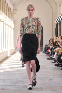 Female model walks a fashion runway in a floral blouse and black draped pencil skirt, audience on either side.
