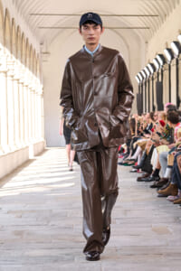 Male model walks a runway in a brown shiny leather suit and navy cap, audience seated along a sunlit arched corridor.