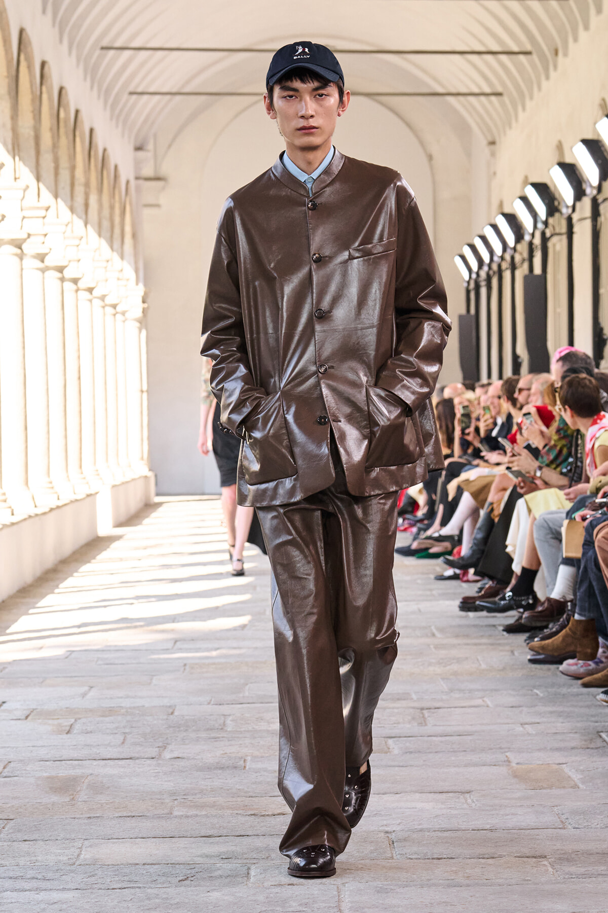Male model walks a runway in a brown shiny leather suit and navy cap, audience seated along a sunlit arched corridor.