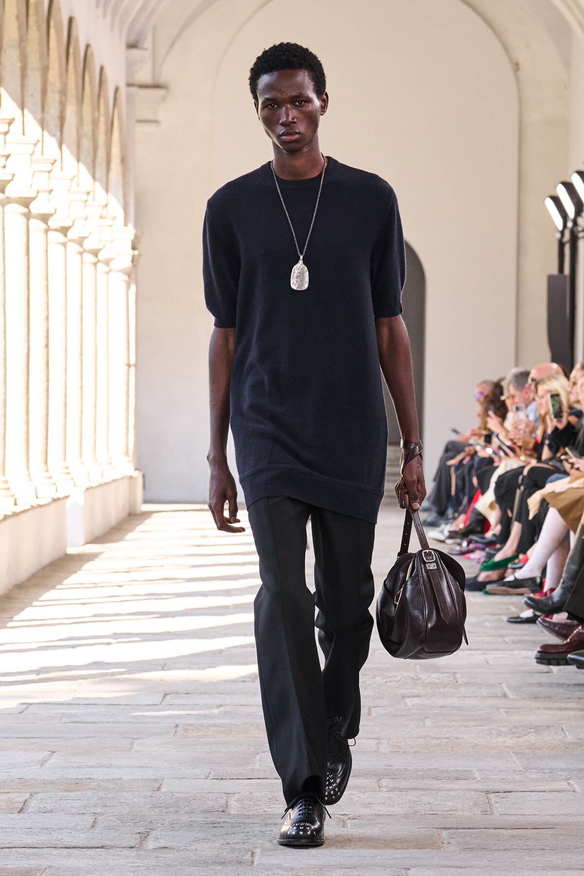 Male model walks a runway in a navy tunic, black trousers, silver pendant necklace, and carries a dark leather duffel bag; seated audience along the side.