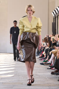Model walks a fashion runway in a pale yellow blouse and brown shiny wrap-skirt, audience watching on both sides.
