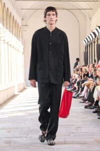 Male model walks a fashion show in a black, minimalist suit with a red tote bag on the runway.