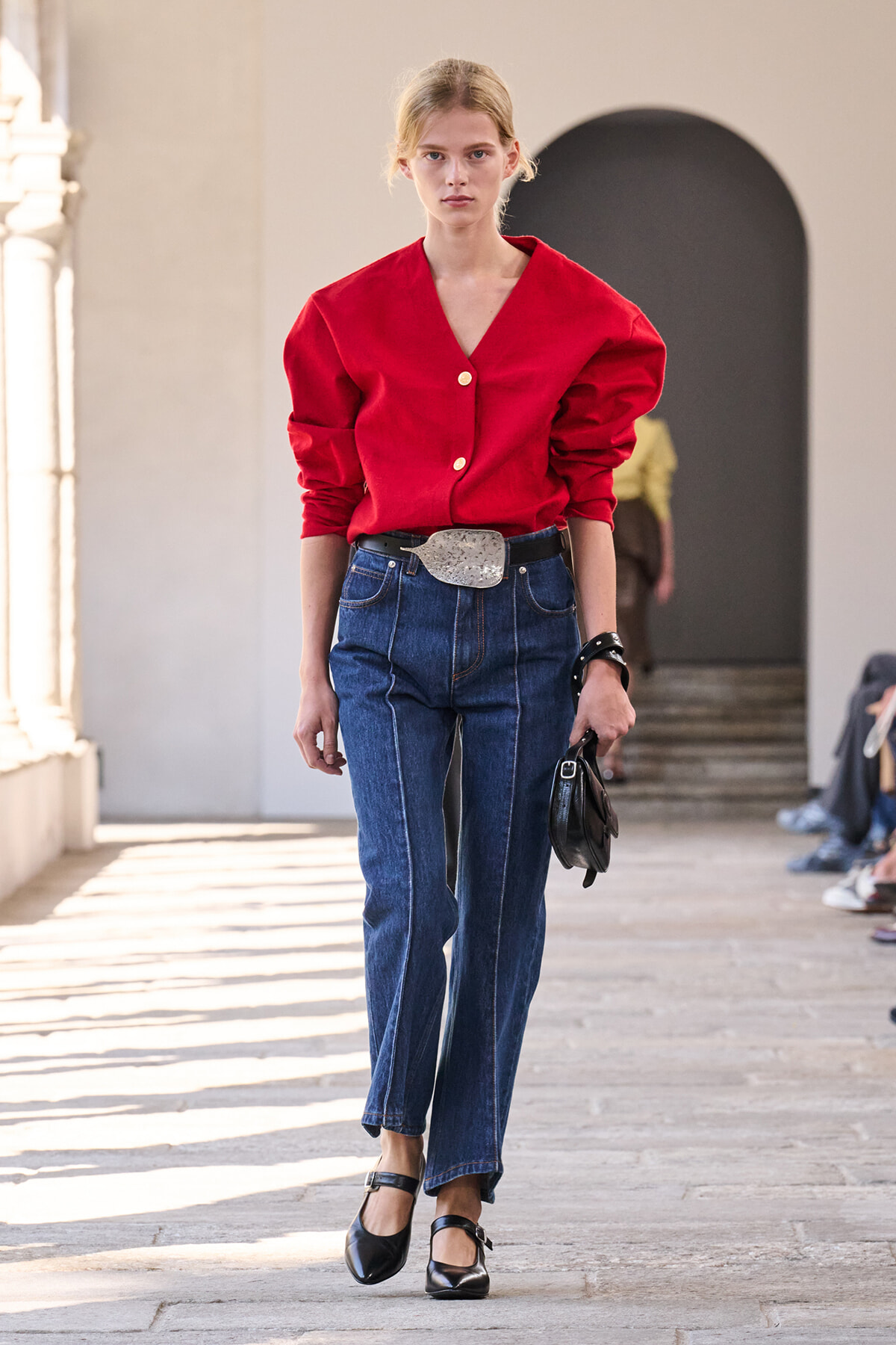 Model walking a runway in a bright red V-neck blouse, high-waisted blue jeans, and a wide silver belt in a sunlit venue.