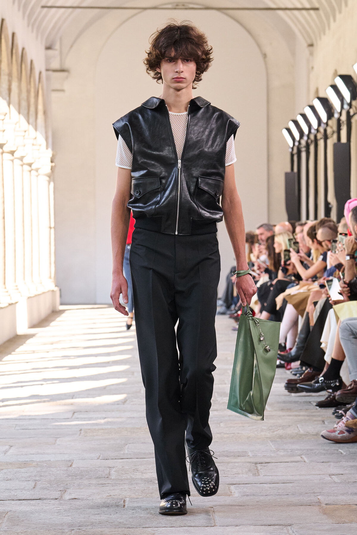 Male model walks the runway in a sleeveless black leather vest over a white mesh tee, black trousers, studded shoes, and carries a green tote.