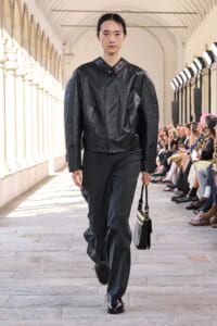 Model walks a runway in a black leather jacket and wide gray trousers, carrying a small two-tone handbag.