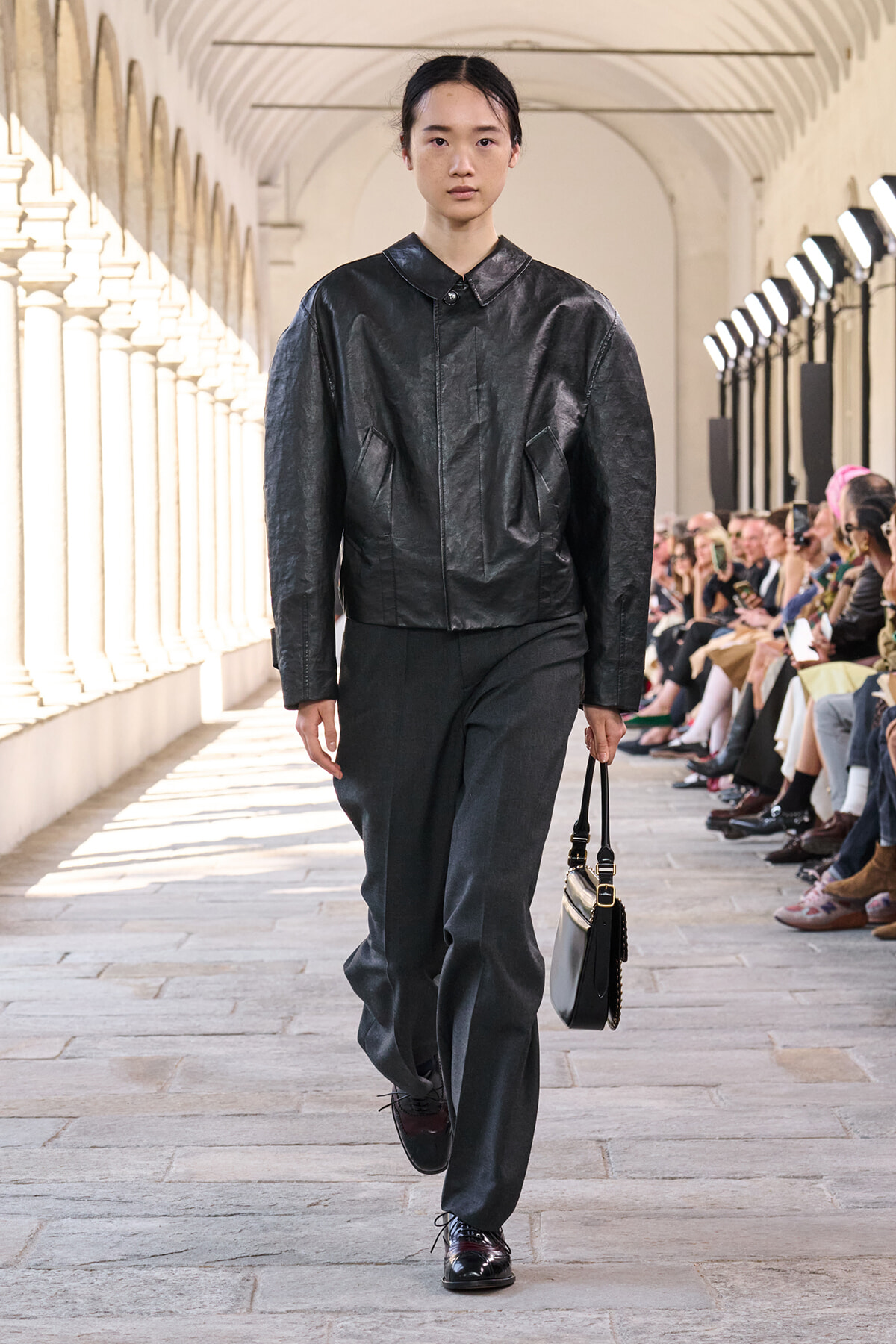 Model walks a runway in a black leather jacket and wide gray trousers, carrying a small two-tone handbag.