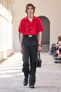 Fashion model walks a runway in a red short-sleeve shirt, dark wide-leg pants, black belt, studded black shoes, and a small brown bag.