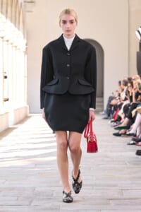 Model walking a runway in a black tailored peplum blazer over a white turtleneck and black skirt, carrying a bright red handbag.