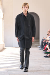 Male model walking a fashion show runway in a knee-length black coat and gray trousers, audience visible on the side.