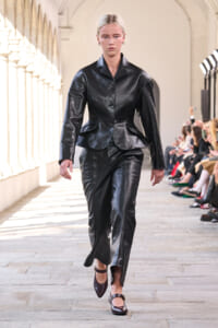 Model walks a runway in a tailored black leather blazer with peplum and matching trousers, in a sunlit arcaded venue.