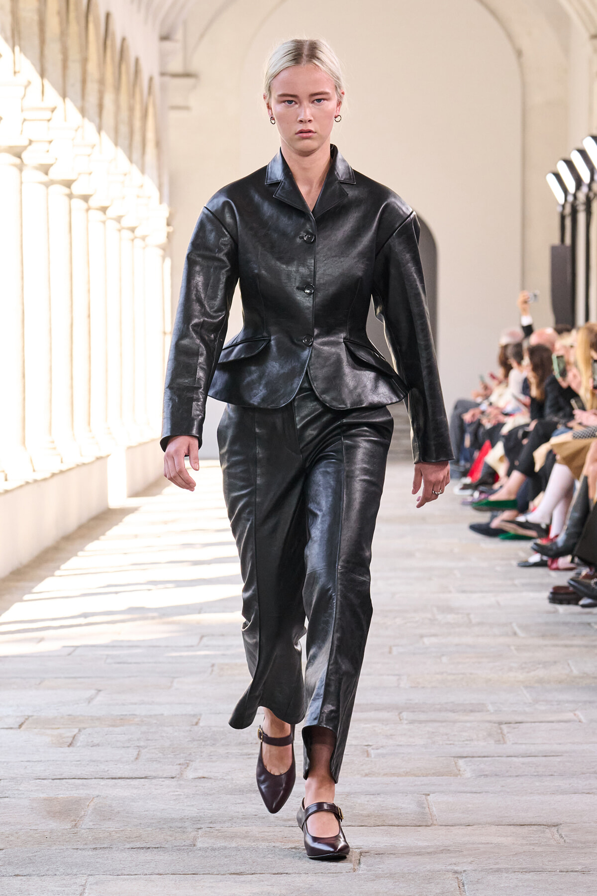 Model walks a runway in a tailored black leather blazer with peplum and matching trousers, in a sunlit arcaded venue.