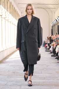 Model walking the runway in a dark gray oversized double-breasted coat, carrying a small brown handbag; audience seated along the sides.