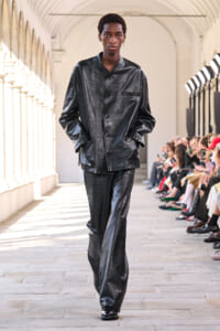 Male model in a black leather suit walking a runway in a bright arched hallway with audience on the sides.