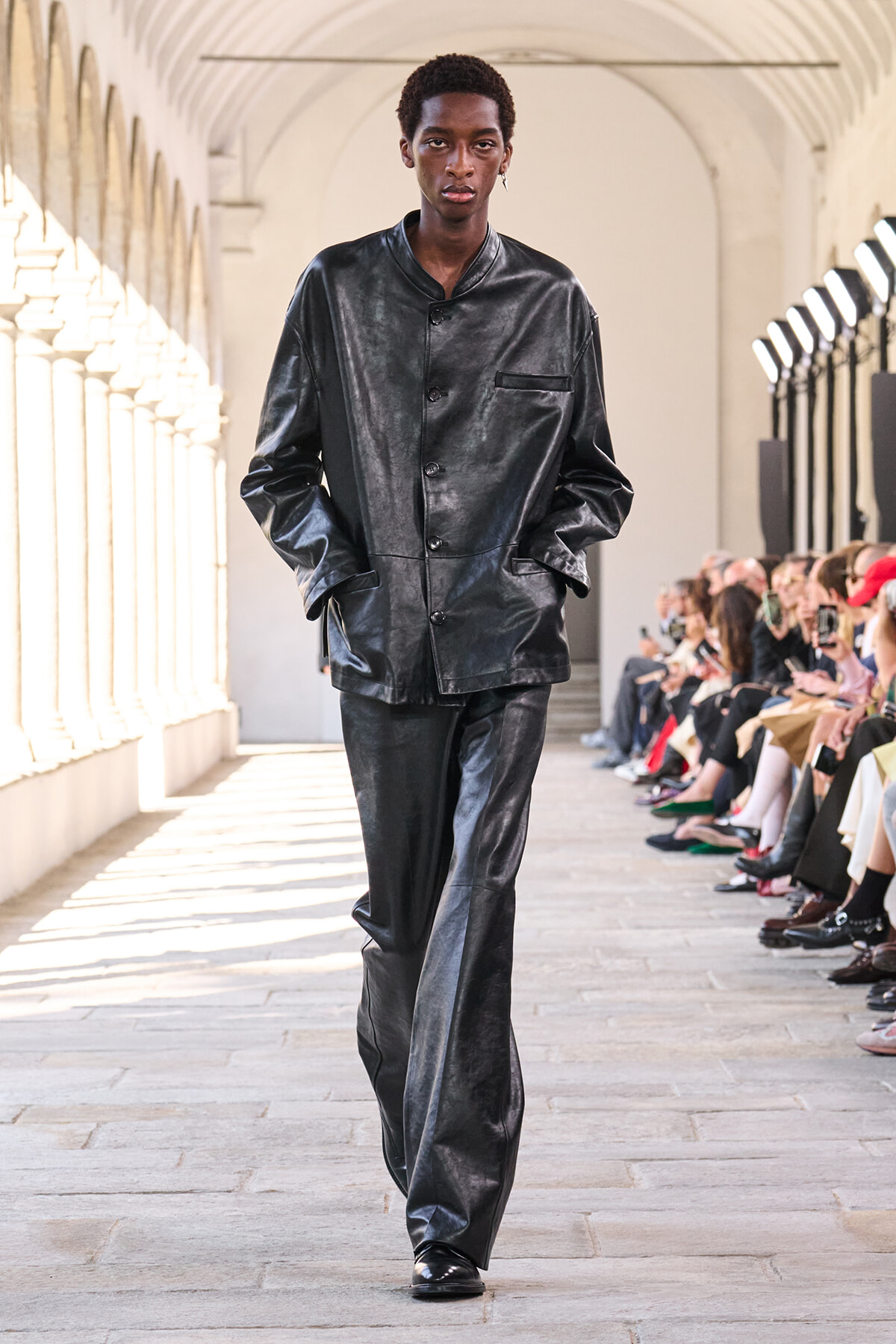 Male model in a black leather suit walking a runway in a bright arched hallway with audience on the sides.
