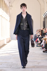 Female model walking a runway in a navy suit with a black leather vest and mesh top, holding a small dark briefcase.