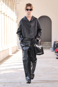 Model walking a runway in a shiny black leather-like suit with sunglasses, carrying a black handbag.