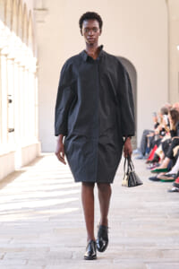 Model walks the runway in a loose black coat and black dress, carrying a small black handbag, with audience seated along the side.