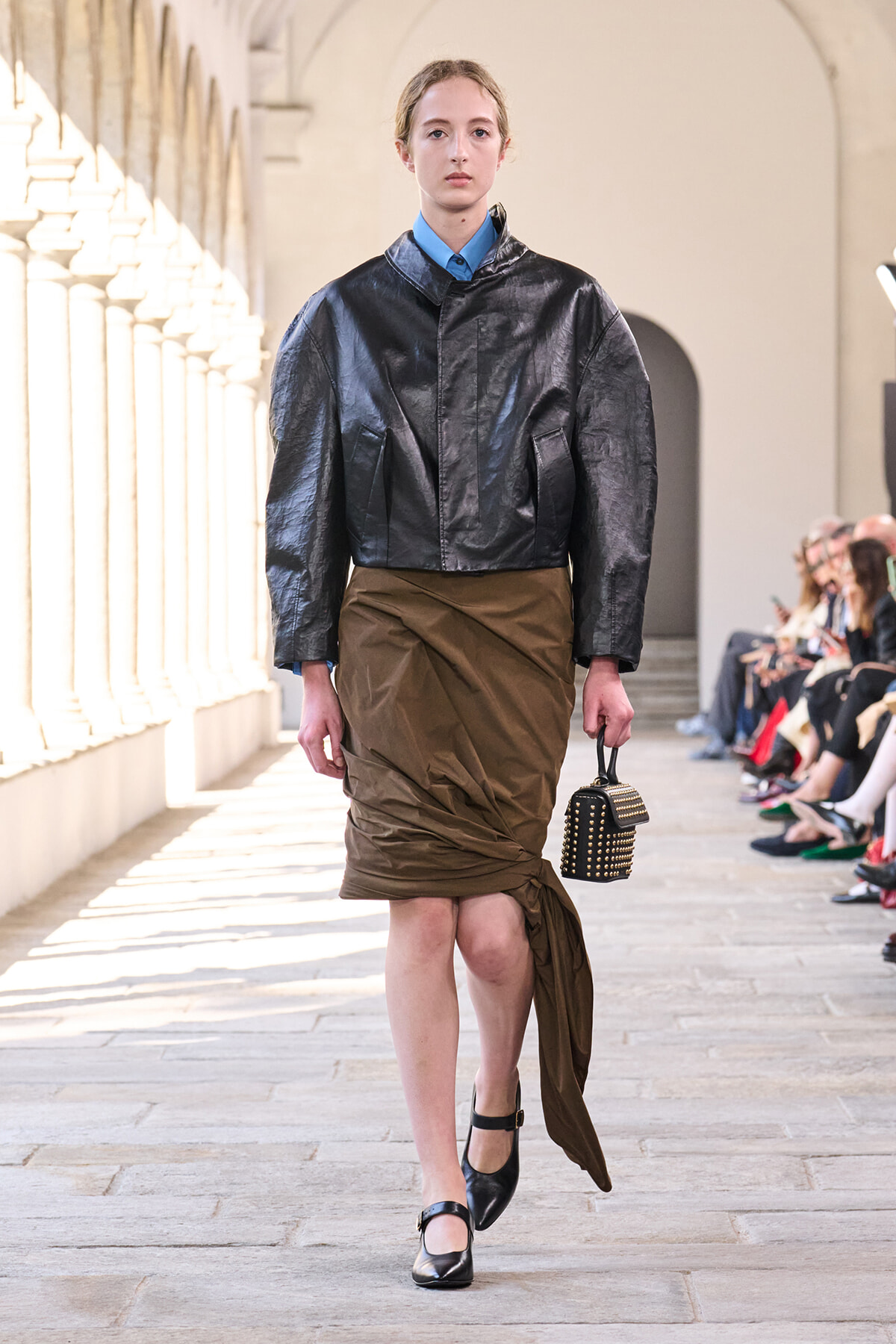 Model on a runway wearing a cropped metallic black jacket, brown draped skirt, blue shirt, black Mary Jane shoes, and a studded handbag.