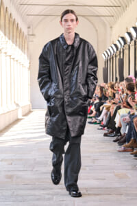 Model walks down a runway wearing a black glossy long coat and dark trousers, surrounded by seated fashion-show spectators in a sunlit arches venue.