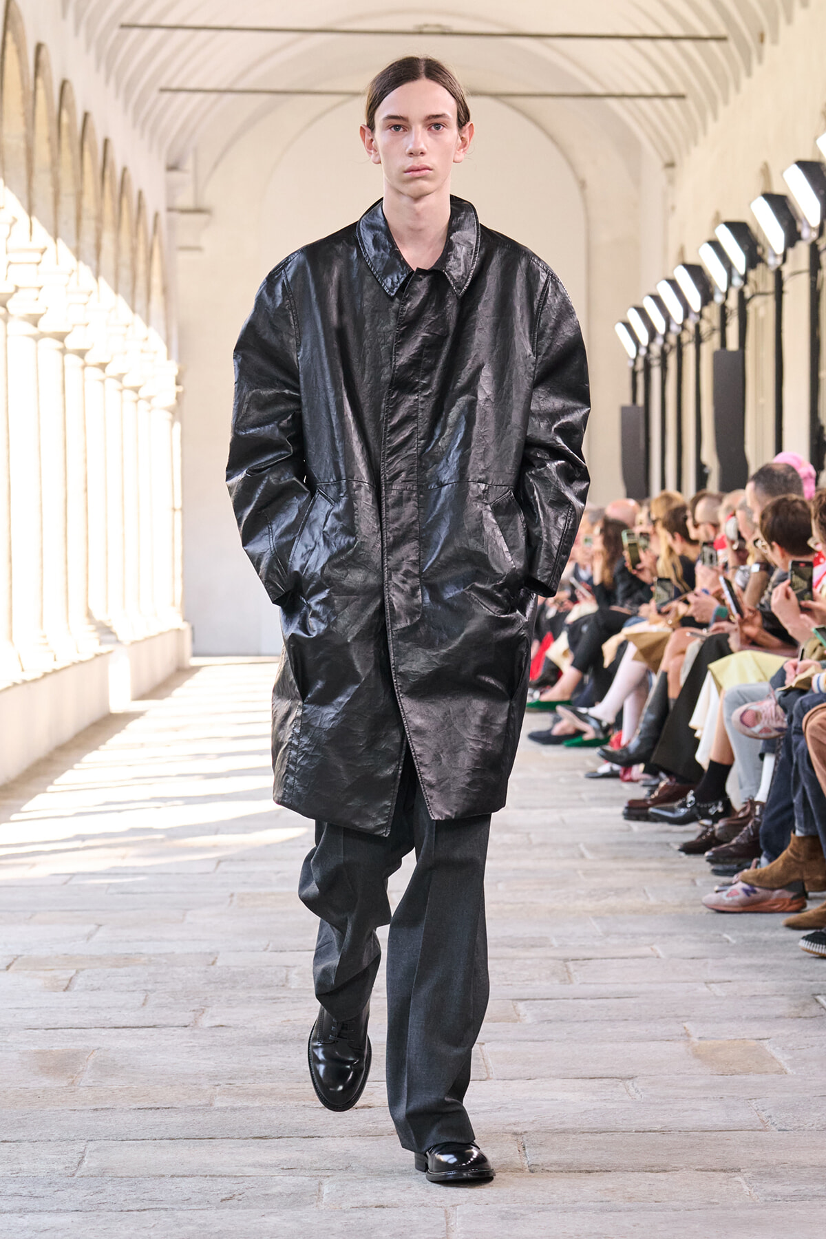 Model walks down a runway wearing a black glossy long coat and dark trousers, surrounded by seated fashion-show spectators in a sunlit arches venue.