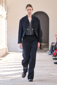 Model walking a runway in a navy suit with a black leather vest and mesh top beneath.